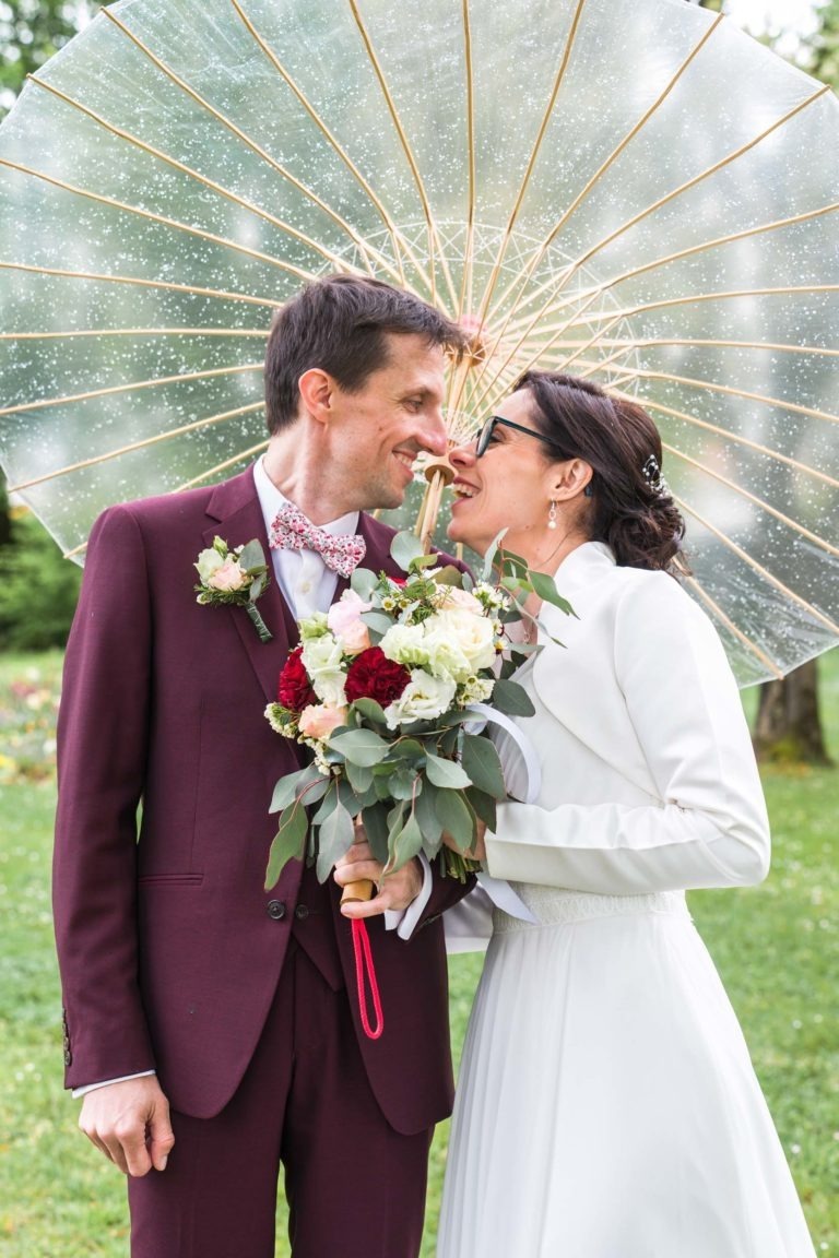 Un couple de mariés rient ensemble sous un parapluie transparent en forme de cœur orné de gouttes de pluie. La mariée, en robe blanche, tient un bouquet de fleurs blanches et rouges, et le marié, en costume bordeaux avec un nœud papillon rose, se penche vers elle. Tous deux ont l'air joyeux au milieu de la verdure pendant leur mariage sous la pluie.