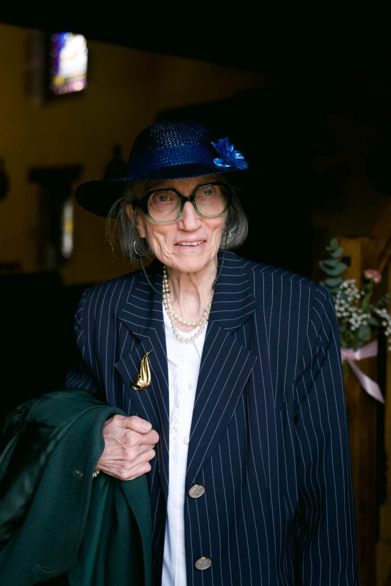 Une femme âgée, vêtue d'un costume à rayures bleu marine, d'une chemise blanche et d'un chapeau bleu marine orné d'une fleur bleue, se tient debout et tient un manteau vert. Elle porte de grandes lunettes à monture noire, un collier de perles et une broche en or en forme d'épingle. L'arrière-plan sombre évoque l'ambiance de "Mariage sous la pluie", avec la lumière des vitraux venant d'en haut.