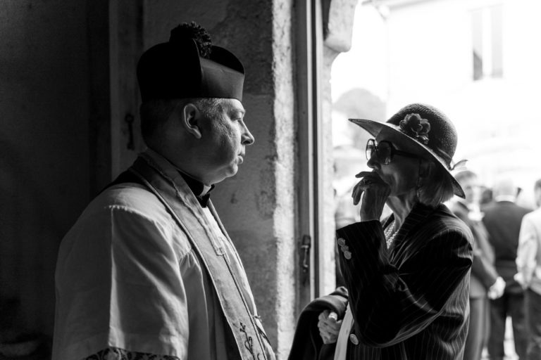 Une photo en noir et blanc montre un prêtre en tenue liturgique en train de discuter avec une femme âgée coiffée d'un chapeau à larges bords orné d'une fleur. Le prêtre regarde la femme qui semble parler, la main levée vers le menton. Derrière eux, un panneau délavé indique « Mariage sous la pluie ».