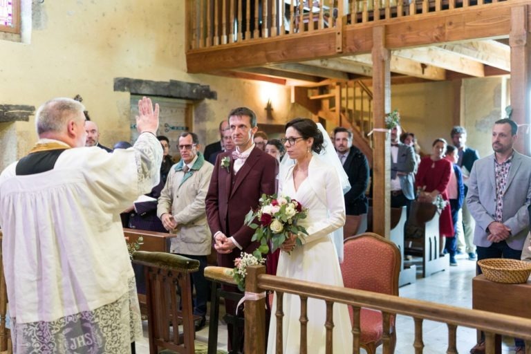 Une mariée et un marié se tiennent ensemble devant l'autel d'une église. Le marié porte un costume marron et la mariée porte une robe de mariée blanche et un voile, tenant un bouquet de fleurs. Devant eux, un prêtre en robe blanche lève la main en signe de bénédiction. Les invités regardent la cérémonie, qui rappelle les scènes de mariage sous la pluie.