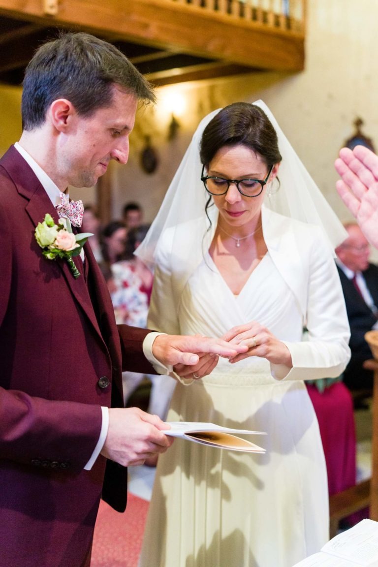 Une mariée et un marié échangent leurs alliances lors de leur cérémonie de mariage. Le marié, vêtu d'un costume bordeaux, place une bague au doigt de la mariée. La mariée est vêtue d'une robe blanche et d'un voile, tenant un papier. Ils sont à l'intérieur avec un balcon en bois et des invités assis en arrière-plan, ce qui laisse présager d'un "mariage sous la pluie" intime.