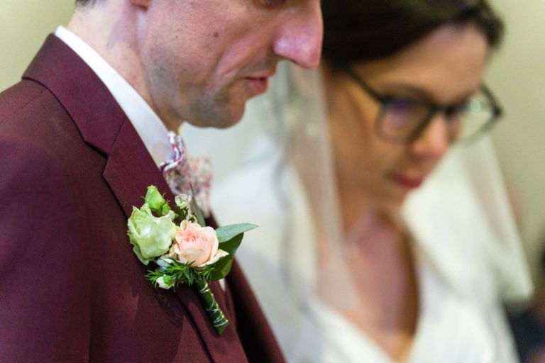 Gros plan d'un couple le jour de leur mariage, peut-être lors d'une cérémonie. Le marié est vu dans un costume bordeaux avec une boutonnière de fleurs blanches et roses sur son revers. La mariée, portant des lunettes et un voile, se tient à côté de lui, légèrement floue. Tous deux regardent vers le bas, rappelant "Mariage sous la pluie".