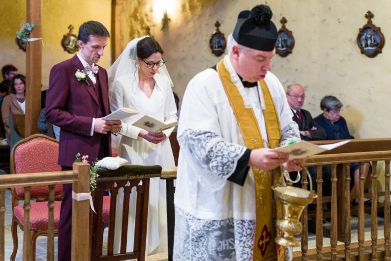 Une mariée en robe blanche et voile se tient à côté d'un marié en costume bordeaux. Tous deux tiennent des brochures et semblent lire lors d'une cérémonie dans une église. Un prêtre en habits ornementés se tient au premier plan, lui aussi en train de lire. Malgré les prévisions de mariage sous la pluie, la cérémonie à l'intérieur est sereine.