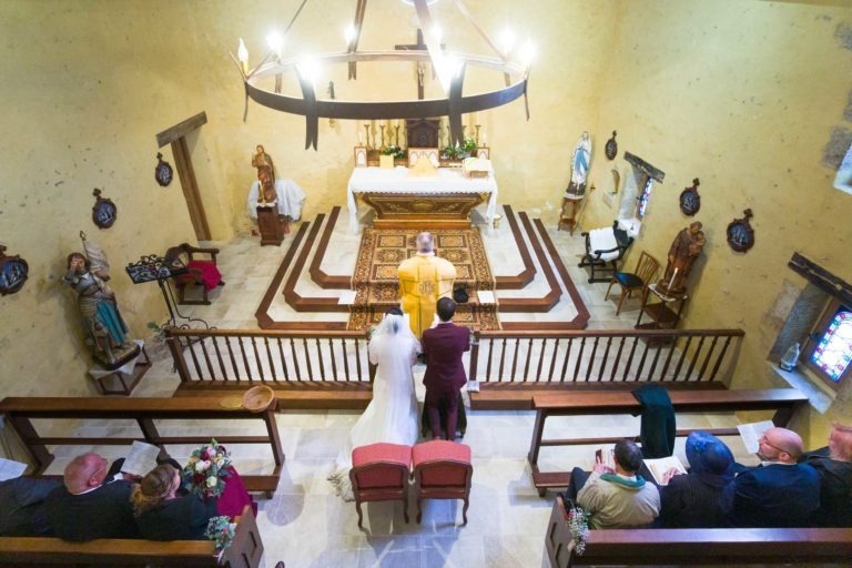 Un couple de mariés se tient devant l'autel lors d'une cérémonie de mariage dans une petite église. L'église, ornée d'un décor simple avec des murs beiges, des bancs en bois et des statues religieuses, fait écho au charme d'un « mariage sous la pluie ». Les invités sont assis tandis que des vitraux baignent la pièce d'une lumière colorée.