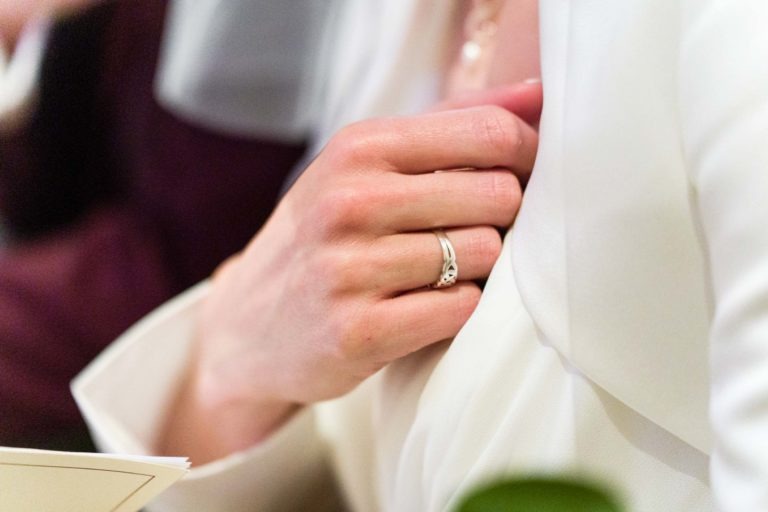 Gros plan d'une personne portant une bague en argent à la main gauche, saisissant délicatement le revers d'un vêtement blanc. L'arrière-plan flou et l'atmosphère formelle évoquent une scène élégante rappelant "Mariage sous la pluie", avec l'accent mis sur la main et la bague, mettant en valeur une élégance subtile.