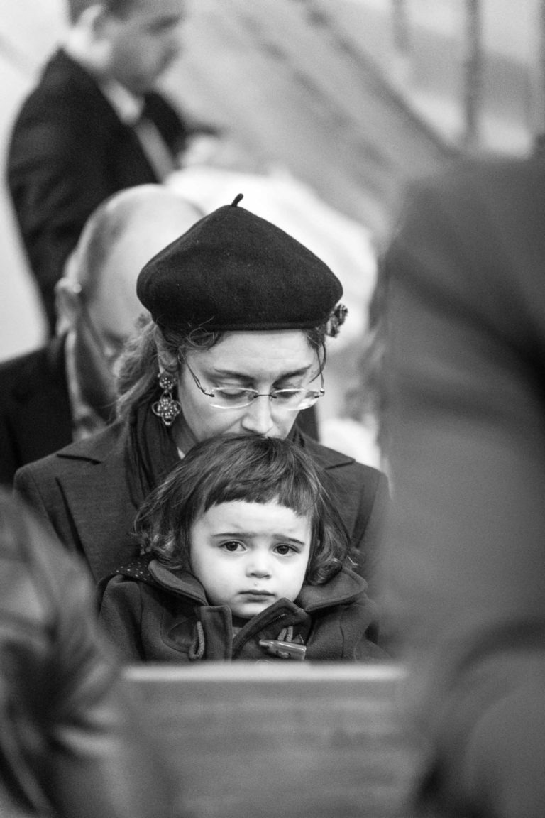 Une image en noir et blanc montre une femme portant un béret et des lunettes, assise juste derrière un jeune enfant aux cheveux courts et foncés. L'enfant semble contemplatif, le regard légèrement baissé. La femme regarde vers le bas, son visage partiellement caché par la tête de l'enfant. D'autres personnages flous rappelant "Mariage sous la pluie" sont à l'arrière-plan.