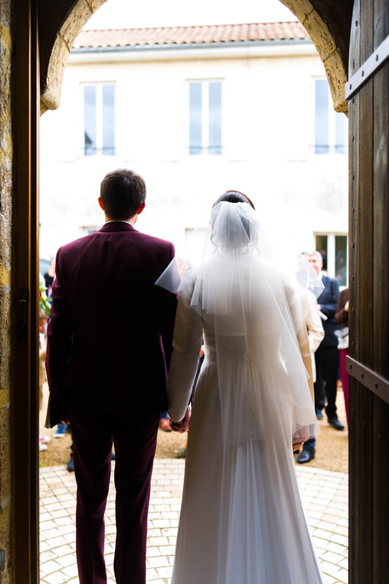 Un couple se tient debout, main dans la main, sous une arche en pierre, dos à la caméra. Le marié porte un costume sombre et la mariée est dans une longue robe blanche avec un voile. Ils sont dehors, où les gens sont rassemblés devant un bâtiment de couleur claire avec de grandes fenêtres. C'est un beau mariage sous la pluie.