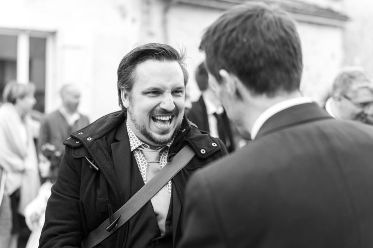 Une photo en noir et blanc montre un homme portant une barbe et un manteau riant avec énergie tout en discutant avec un autre homme en costume, dos à la caméra. La scène, qui évoque "Mariage sous la pluie", semble se dérouler lors d'une réunion sociale, avec des silhouettes floues et un bâtiment en arrière-plan.