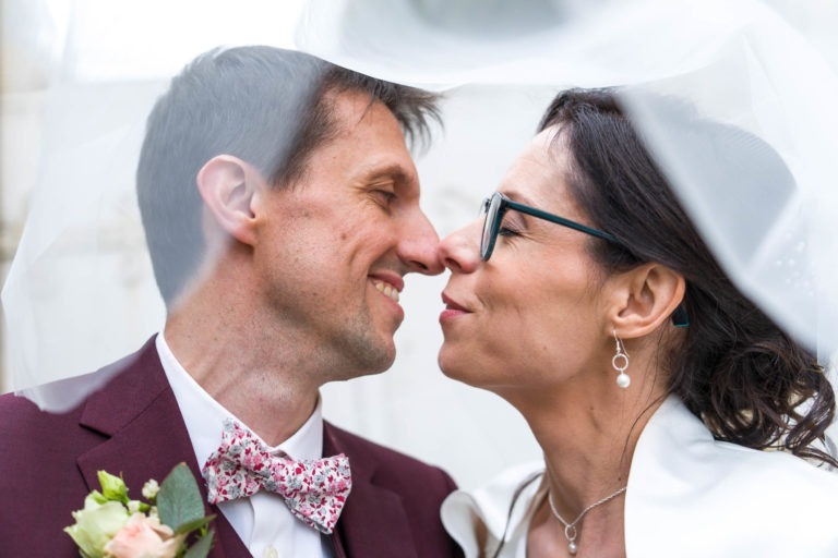 Un couple de mariés se tient près l'un de l'autre, le nez en contact, sous un voile blanc. Le marié porte un costume bordeaux avec un nœud papillon fleuri et une boutonnière. La mariée porte des lunettes, des boucles d'oreilles et une robe blanche, souriant doucement. Tous deux partagent un moment intime le jour de leur mariage, rappelant le mariage sous la pluie.