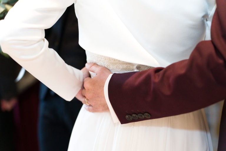 Une personne en robe de mariée blanche est vue de dos, les mains posées sur les hanches. Un autre individu portant une veste marron se tient à proximité, sa main posée délicatement sur le bas du dos de la personne, créant un geste intime et solidaire. L'arrière-plan est flou, rappelant un mariage sous la pluie.