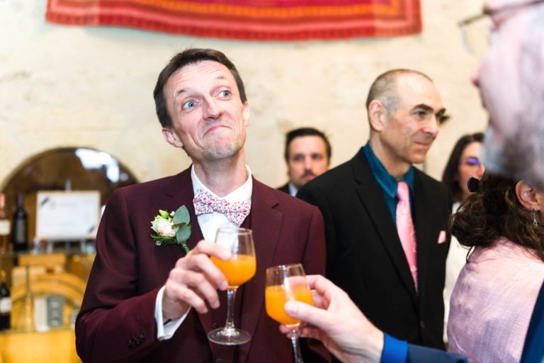 Un homme en costume marron et nœud papillon fleuri tient un verre de jus d'orange, faisant un visage expressif. Il a une boutonnière sur son revers de veste, célébrant probablement un mariage sous la pluie. Entouré de personnes en tenue de soirée dans une pièce avec une tapisserie sur le mur, d'autres autour de lui tiennent également des verres, participant probablement à un toast.