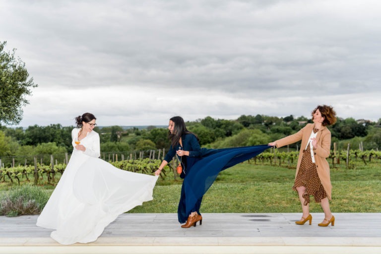 Trois femmes se tiennent dehors sur un chemin avec un vignoble en arrière-plan. La femme en robe blanche à gauche étend son bras pour tenir la robe de la femme du milieu, qui porte une tenue bleu foncé. La femme à droite, en tenue marron, tient la robe de la femme du milieu, toutes souriantes comme si elles célébraient « Mariage sous la pluie ».