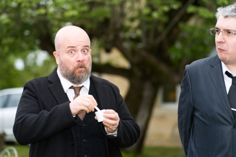 Deux hommes se tiennent dehors, avec un arbre vert luxuriant en arrière-plan. L'homme de gauche, avec une tête chauve et une barbe, a une expression surprise alors qu'il regarde directement la caméra tout en tenant une petite tasse blanche et une cuillère. L'homme de droite, portant des lunettes et un costume, regarde sur le côté, rappelant une scène de "Mariage sous la pluie".