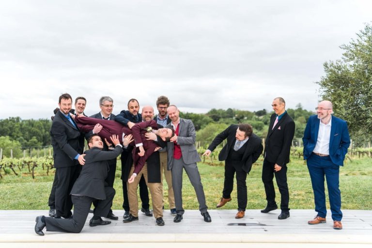 Un groupe d'hommes en costume, réunis à l'extérieur, tiennent de manière ludique un homme en costume marron à l'horizontale. Ils semblent célébrer et rire de ce joyeux mariage sous la pluie, certains tendant la main pour soutenir l'homme ou posant avec humour. L'arrière-plan présente une végétation verte et un ciel nuageux.