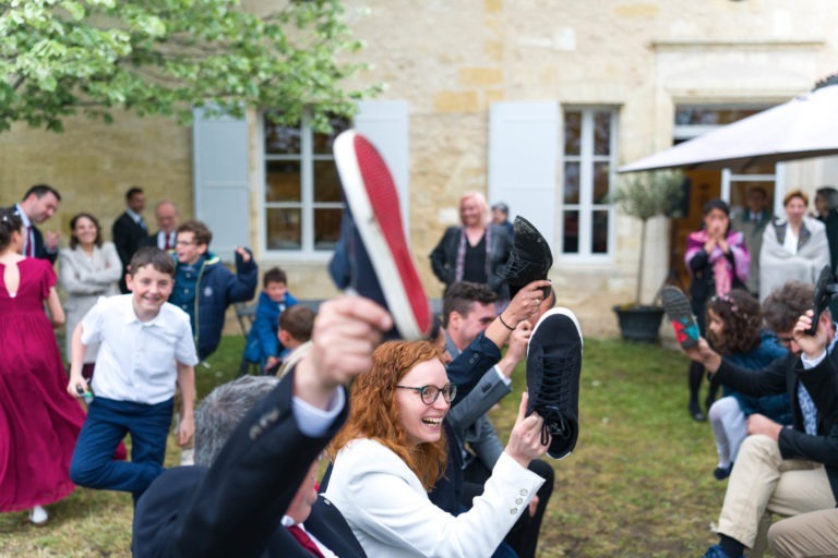 Un groupe de personnes, composé d’enfants et d’adultes, se livre à une activité de plein air animée. Certains individus brandissent des chaussures. Au premier plan, une femme aux cheveux roux bouclés et portant des lunettes s’amuse visiblement. Le décor semble être un jardin avec un bâtiment en pierre à l’arrière-plan, rappelant un « Mariage sous la pluie ».