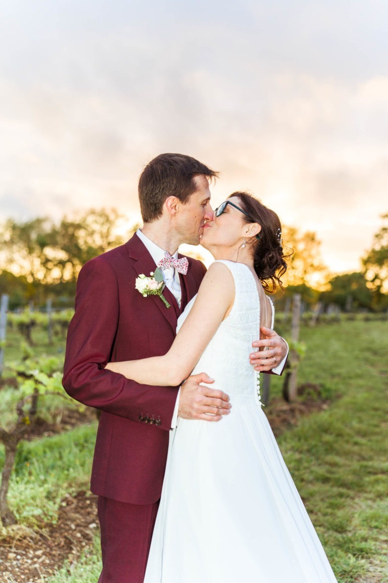 Un couple de mariés s'embrassent en plein air au coucher du soleil. La mariée porte une robe blanche et le marié est habillé d'un costume bordeaux avec un nœud papillon fleuri. Ils se tiennent sur une pelouse avec un vignoble en arrière-plan, baigné d'une lumière chaude et dorée, un contraste parfait avec leur précédent mariage sous la pluie.