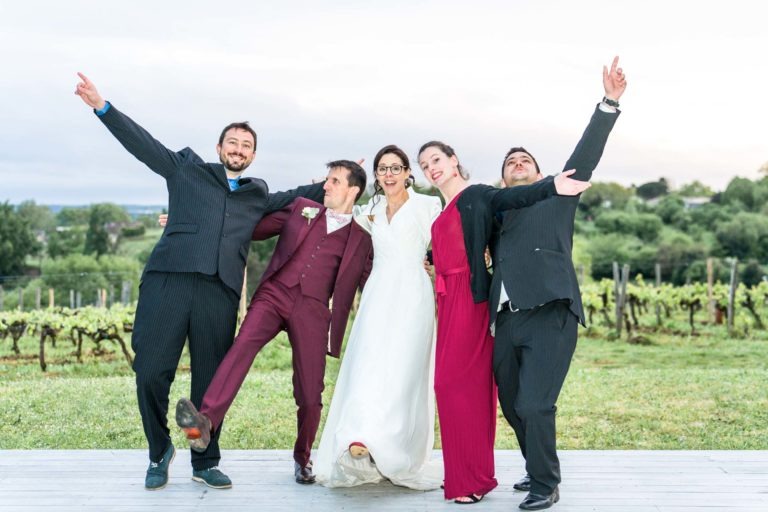 Cinq personnes se tiennent debout dans une pose festive sur une terrasse en bois devant un vignoble, rappelant un mariage sous la pluie. Le groupe comprend deux femmes en robe (une blanche, une rouge) et trois hommes en costume (deux noirs, un marron). Les bras levés, ils sourient tous et ont l'air joyeux sur fond de verdure luxuriante et de vignes.