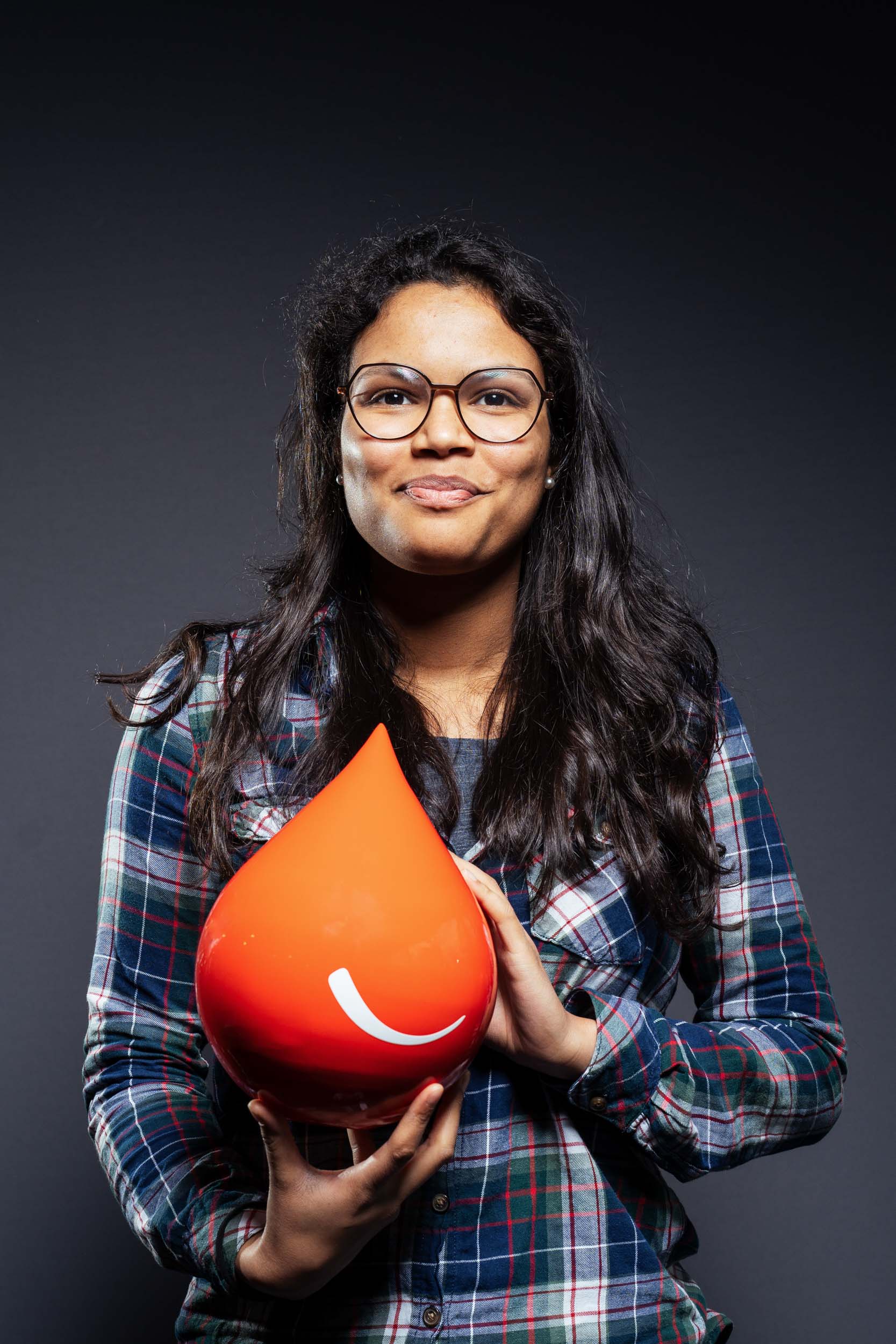 Une femme aux cheveux longs et noirs, portant des lunettes et une chemise à carreaux, se tient debout sur un fond sombre. Elle tient un gros objet rouge vif en forme de goutte avec un visage souriant blanc dessus. Pour ceux qui recherchent des portraits professionnels à Bordeaux, son léger sourire et son regard direct ont un impact.