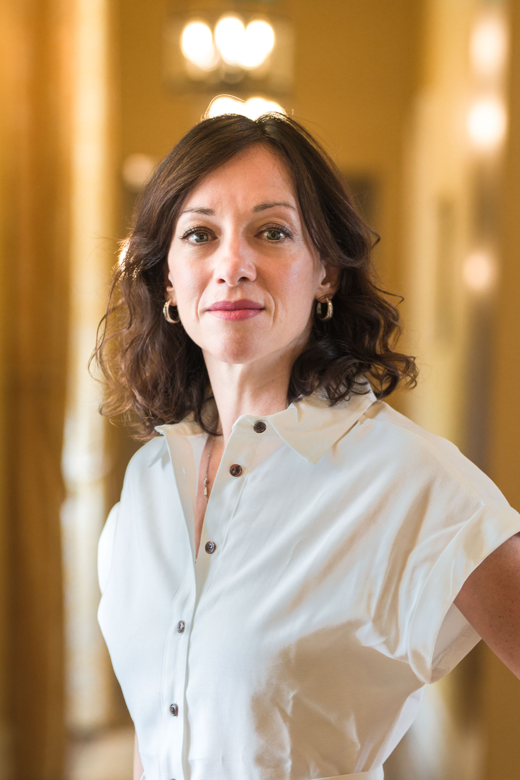 Une femme aux cheveux bruns bouclés jusqu'aux épaules se tient debout dans un couloir bien éclairé. Elle porte une chemise blanche à manches courtes boutonnée et de petites boucles d'oreilles créoles. Avec une expression confiante et les mains posées sur ses hanches, elle incarne l'apparence soignée souvent recherchée dans les portraits professionnels à Bordeaux, tandis que la lumière des fenêtres crée une lueur chaleureuse et invitante en arrière-plan.