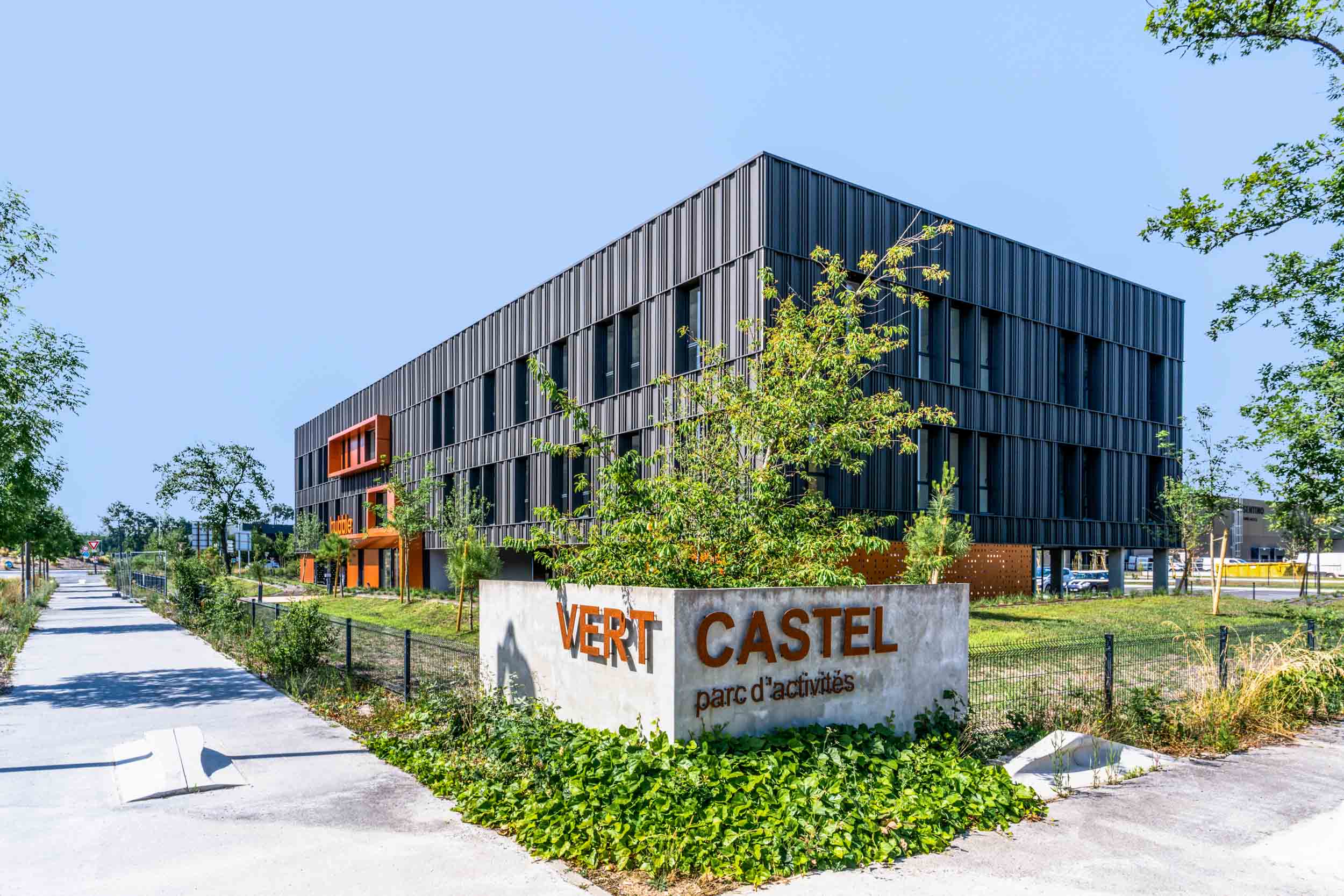 Un bâtiment moderne aux panneaux noirs du parc d'activités Vert Castel est représenté, avec un design anguleux et net avec des reflets orange. Photographié par un talentueux photographe d'architecture, des arbres et des arbustes verts entourent la structure tandis qu'un panneau en béton indique « VERT CASTEL parc d'activités ».