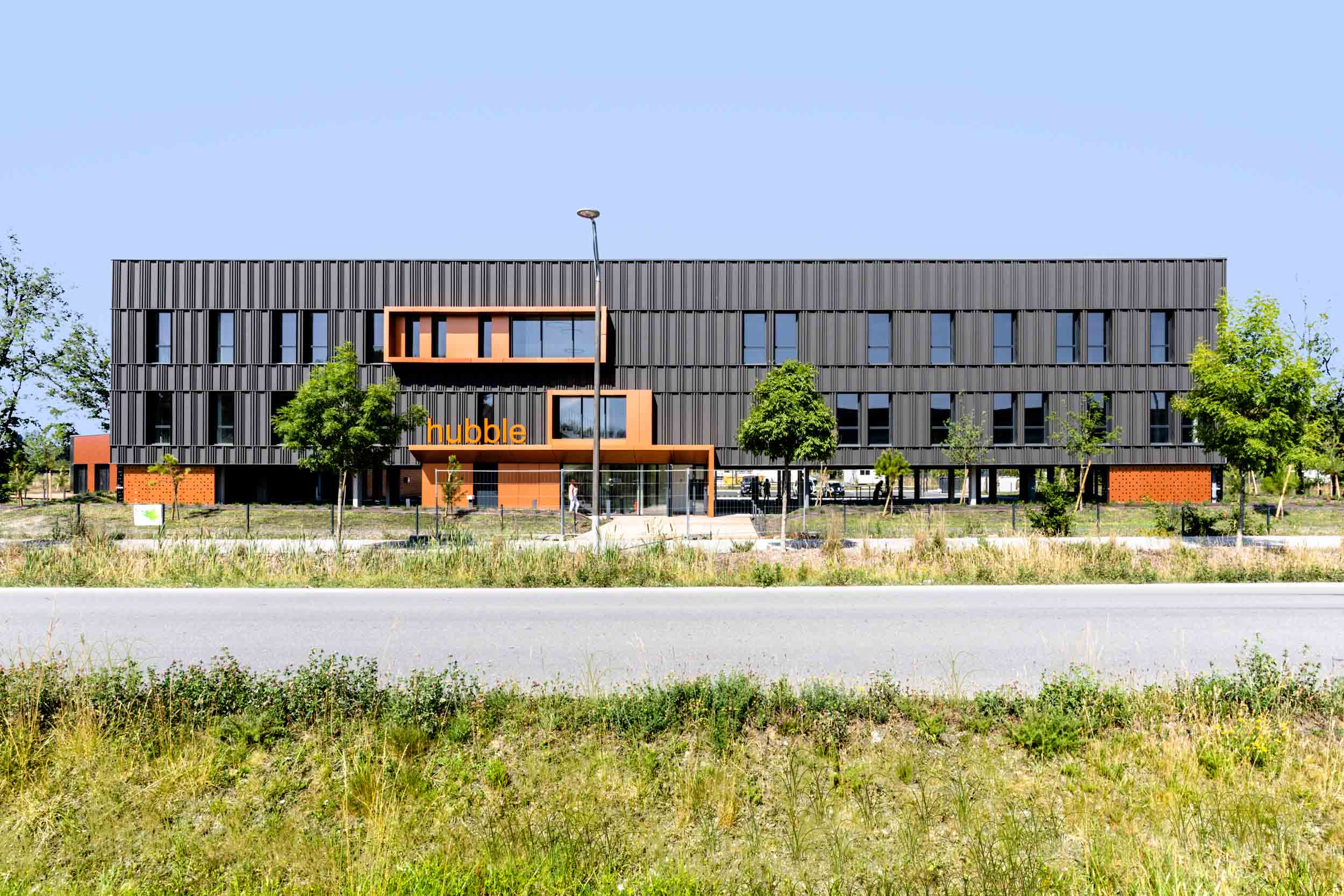 Un bâtiment moderne de trois étages avec une façade gris foncé et des touches d'orange, étiqueté "hubbell" à l'entrée. Il est entouré d'un espace paysager soigné avec des arbres et de l'herbe, et une route passe devant le bâtiment. Le ciel est clair et bleu, offrant la journée parfaite pour la photographie d'architecture.