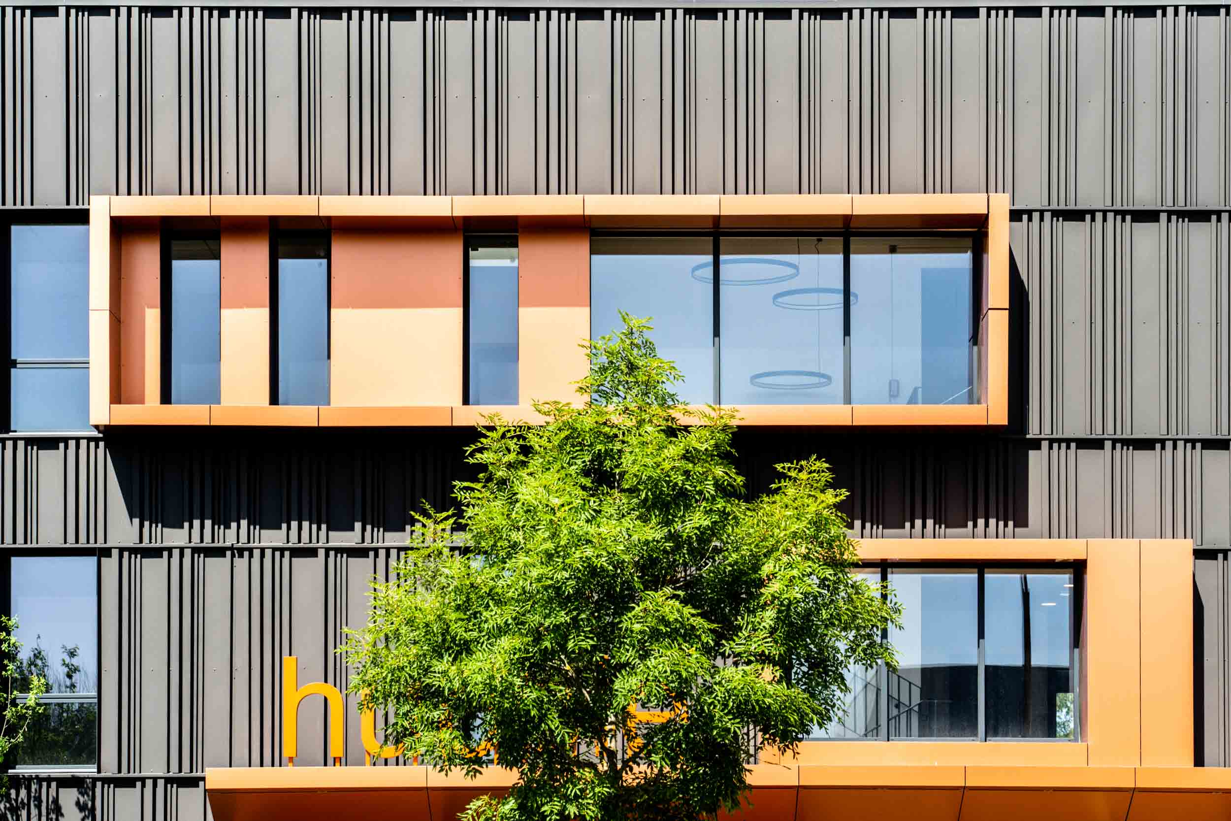 Une façade de bâtiment moderne aux lignes métalliques verticales et horizontales avec une fenêtre rectangulaire proéminente dans des tons de marron et de beige, capturée par un photographe d'architecture talentueux. En dessous, partiellement masquée par un arbre feuillu vert, la signalisation jaune indique « hu ». Des plafonniers circulaires sont visibles à l'intérieur à travers la fenêtre.