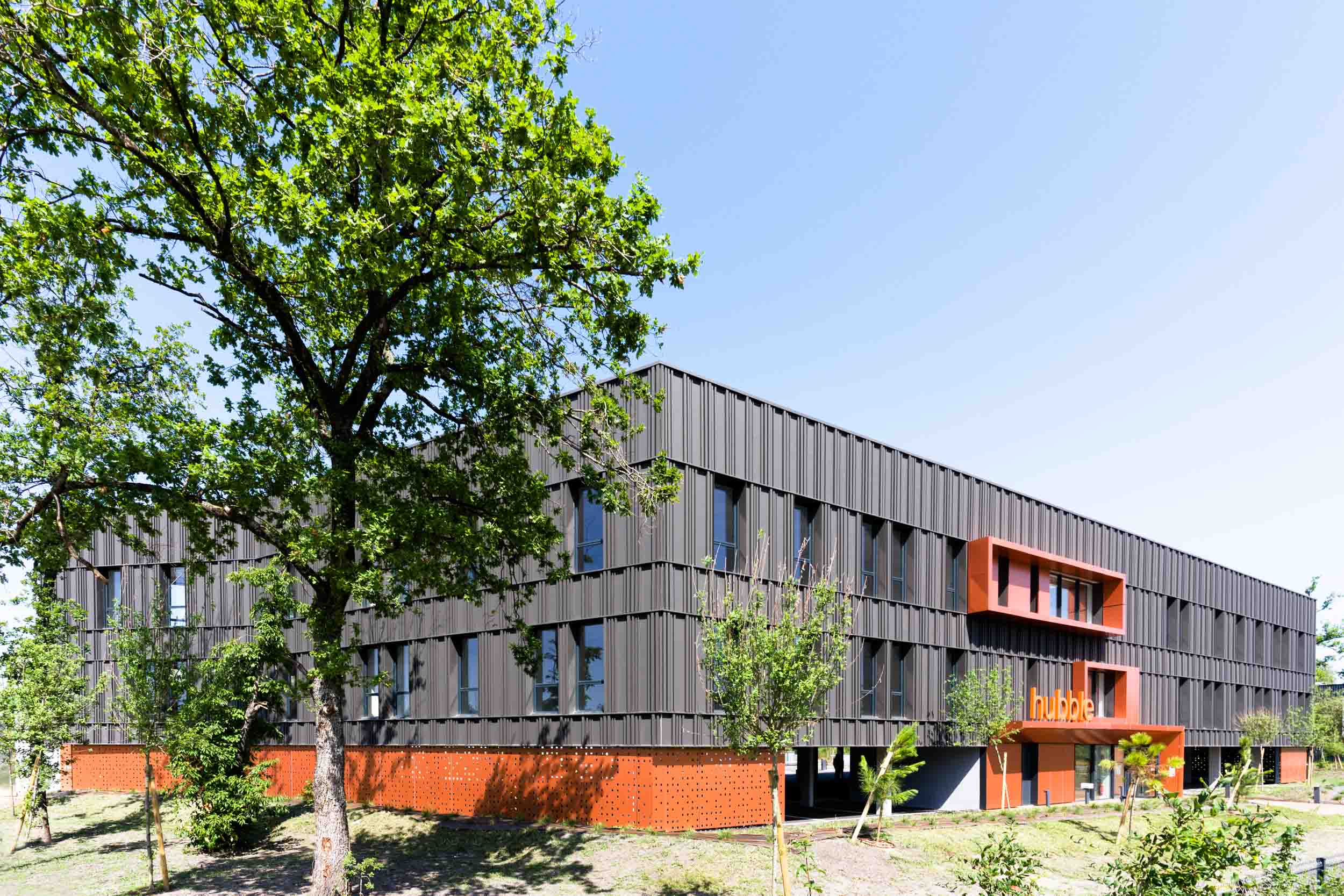 Un bâtiment moderne aux panneaux noirs, aux grandes fenêtres et aux touches de rouge sur la façade, se dresse au milieu d'une pelouse verte parsemée de jeunes arbres. La structure compte deux étages et le mot « hub » est visible en lettres orange en gras. Le ciel bleu clair offre une toile de fond lumineuse à cet exemple de photographie d'architecture.