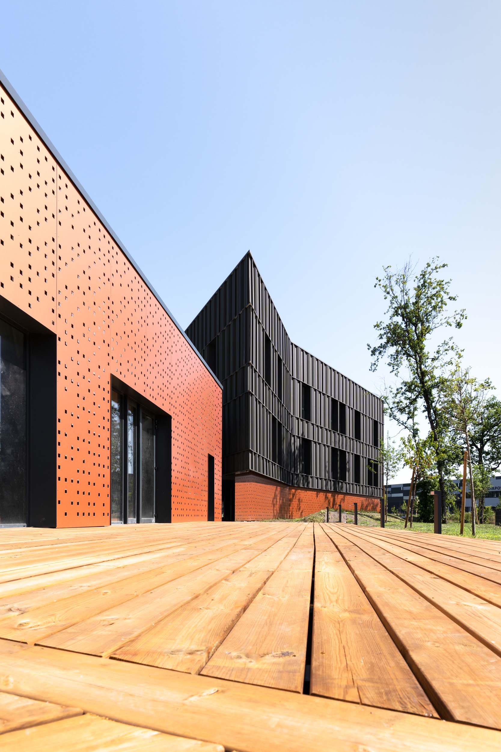 Bâtiment moderne avec une grande terrasse en bois au premier plan. La structure est dotée de panneaux métalliques rouges et noirs avec des motifs perforés sur les panneaux rouges. La photographie d'architecture capture des lignes angulaires, de grandes fenêtres en verre et des arbres verts environnants. Le ciel au-dessus est clair et bleu.