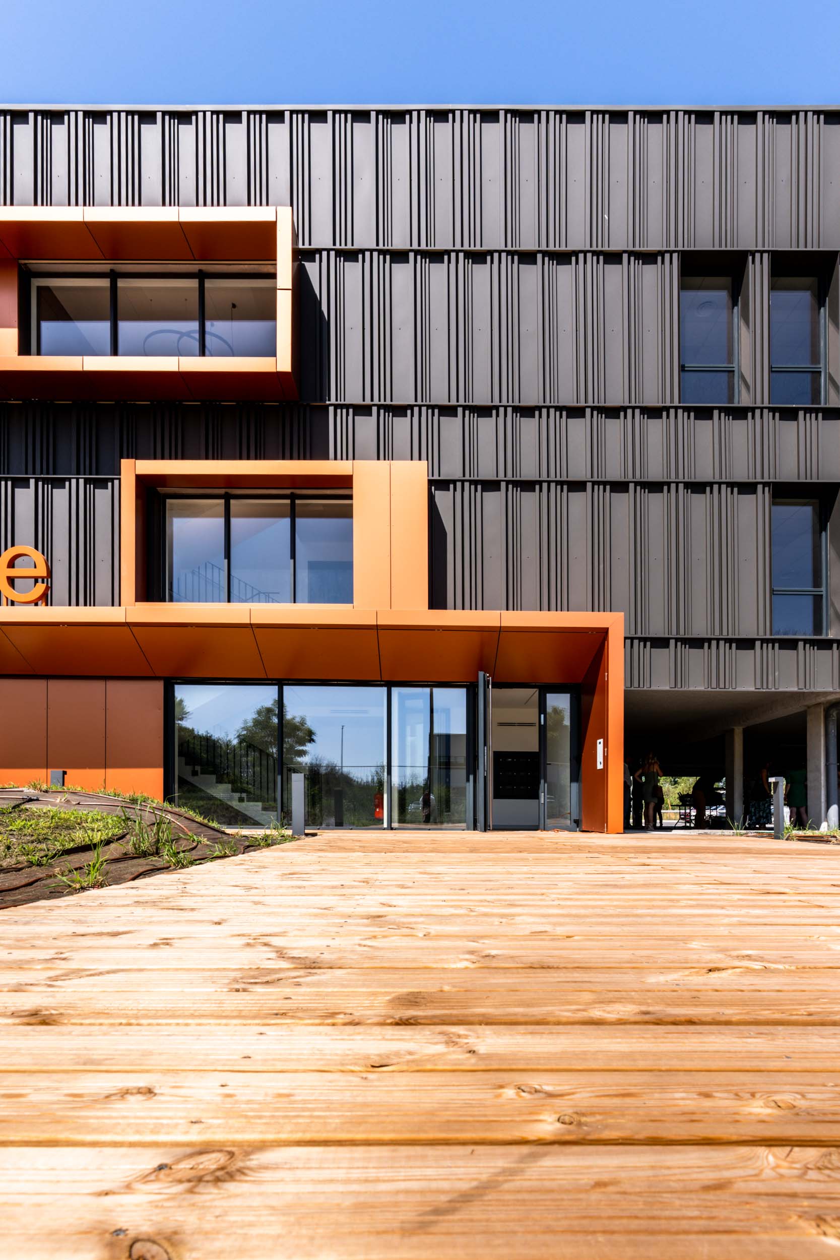 Façade d'un bâtiment moderne avec des panneaux verticaux gris foncé et de grandes fenêtres et une entrée à cadre orange. L'entrée est dotée de grandes portes vitrées, complétées par un chemin en bois qui y mène. Sous le ciel bleu clair d'une journée ensoleillée, un peu de verdure est visible sur la gauche - un cliché parfait pour tout photographe d'architecture.