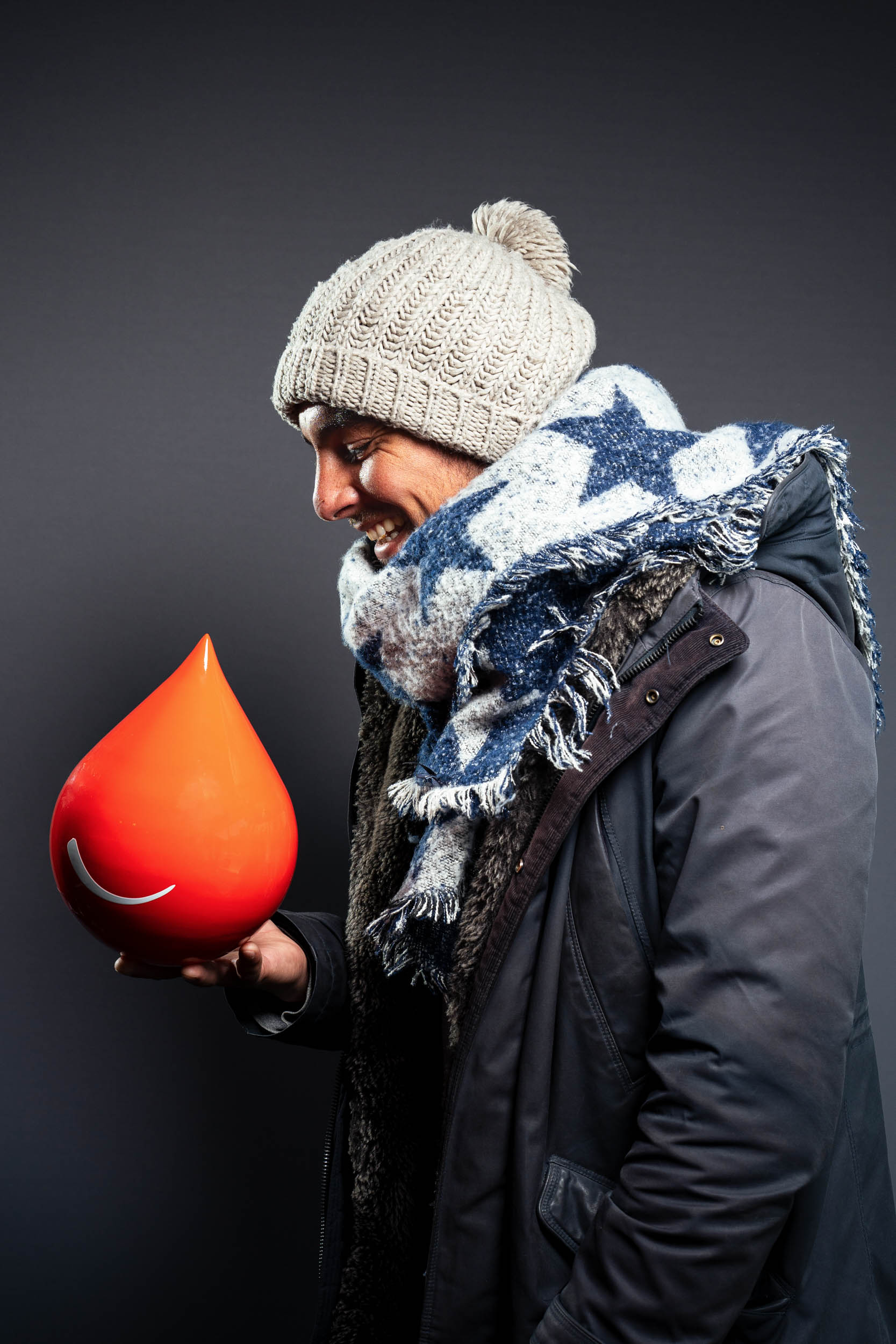 Une personne souriante portant un bonnet tricoté, une écharpe bleue et blanche et un manteau sombre se tient debout sur un fond gris. Dans cet exemple de portraits professionnels à Bordeaux, ils tiennent un grand objet rouge brillant en forme de larme avec une ligne courbe blanche ressemblant à un sourire. La personne regarde l'objet avec joie.