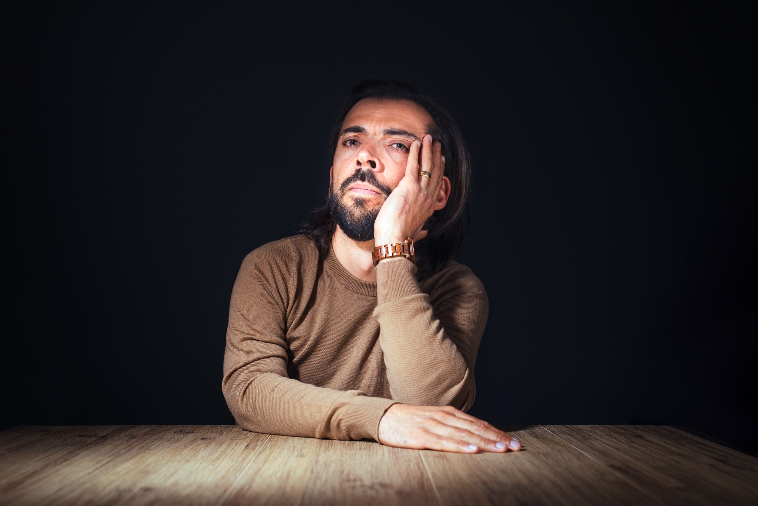 Un homme aux cheveux et à la barbe noirs, vêtu d'une chemise marron à manches longues et d'une montre en or, est assis à une table en bois sur un fond sombre. Il pose sa tête sur sa main, l'air contemplatif, le coude sur la table et l'autre bras tendu vers l'avant - un sujet idéal pour des portraits professionnels à Bordeaux.