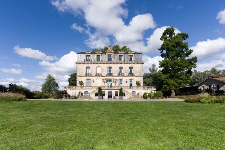 Un grand bâtiment en pierre de trois étages avec des fenêtres cintrées au dernier étage, sur fond de ciel bleu parsemé de nuages. Le bâtiment a un toit bordé d'arbres et est entouré d'une végétation luxuriante et d'une pelouse bien entretenue. Un large escalier mène à l'entrée, parfaite pour accueillir la soirée annuelle du groupe Betclic. Une structure plus petite est visible à droite.