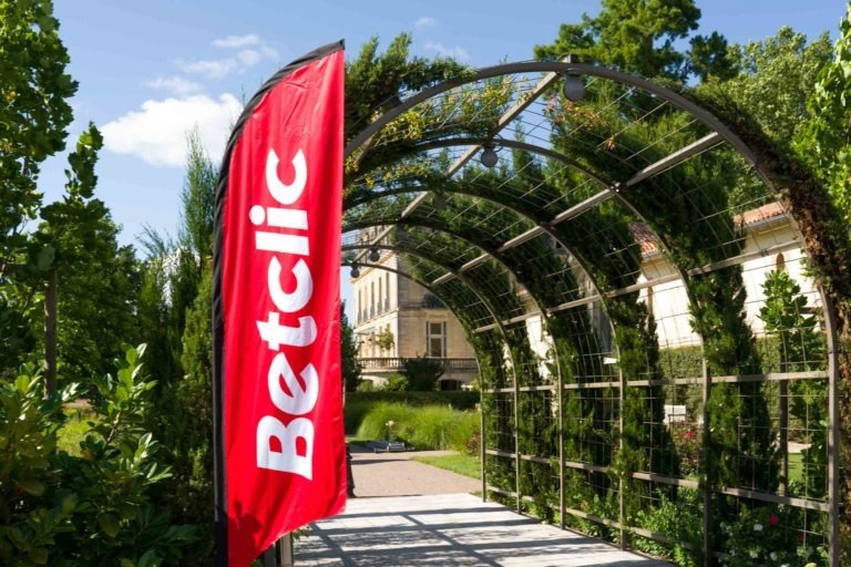 Une grande bannière rouge avec le logo blanc « Betclic » trône près de l'entrée d'une allée de jardin avec une structure en arche métallique recouverte de verdure. L'arrière-plan montre une journée ensoleillée avec un bâtiment partiellement visible à travers les arbres, plantant le décor pour la Soirée annuelle du Groupe Betclic.