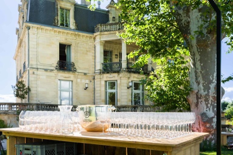 Une table en bois est dressée à l'extérieur avec de nombreux verres à vin vides et un grand seau à glace en argent, prêts pour la soirée annuelle du groupe Betclic. La table est placée devant un élégant bâtiment en pierre claire à plusieurs étages avec des balcons et des détails complexes. Une végétation luxuriante et un grand arbre fournissent de l'ombre et de l'ambiance en arrière-plan.