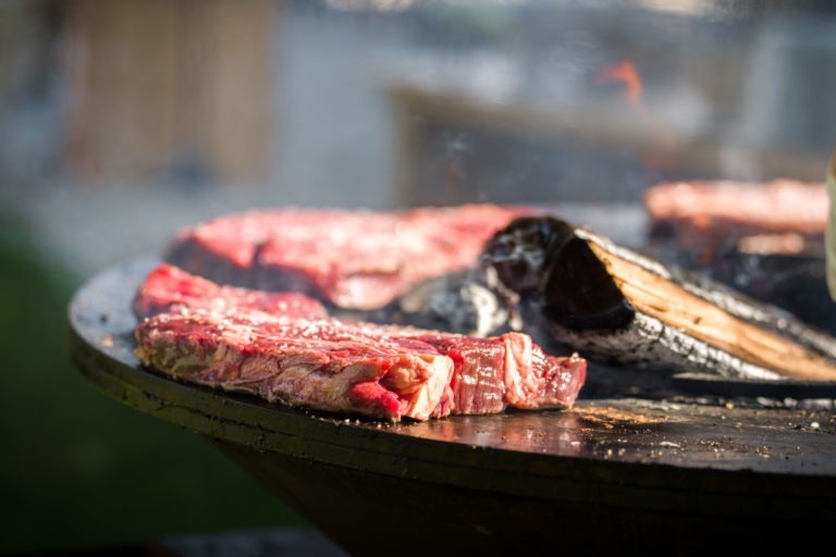 Des steaks grésillants sur un gril extérieur avec des marbrures et des marques de gril visibles, libérant une fumée aromatique à la Soirée annuelle du Groupe Betclic. Un morceau de bois est carbonisé à proximité, mettant en valeur l'environnement de cuisson rustique. L'arrière-plan est flou, soulignant l'accent mis sur la cuisson de la viande.