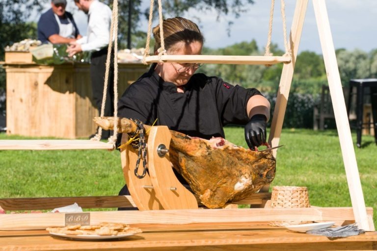 Lors de la Soirée annuelle du Groupe Betclic, un chef vêtu d'un uniforme noir et de gants découpe avec brio la viande d'un gros jambon cru monté sur un support en bois à l'extérieur. En arrière-plan, deux autres personnes en tablier préparent de la nourriture à une table en bois. Le décor est une zone herbeuse avec des arbres et une rivière visible au loin.