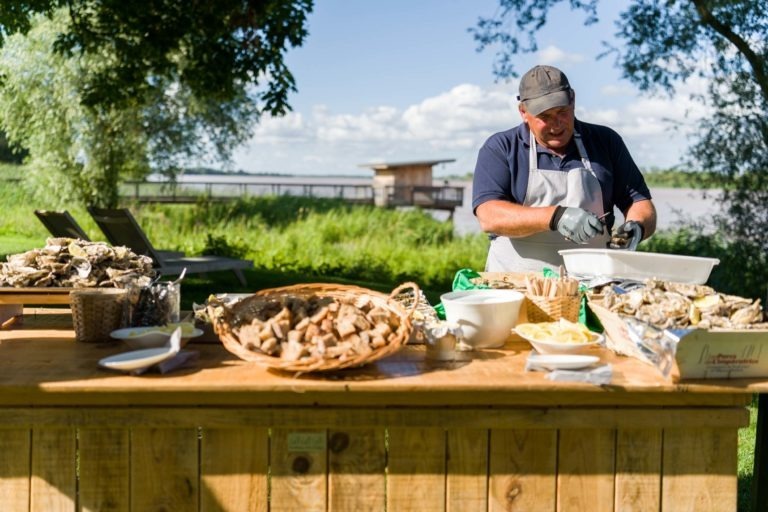 Un homme se tient debout à l'extérieur derrière une table en bois et prépare des huîtres lors de la soirée annuelle du groupe Betclic. Il porte une chemise sombre, un tablier, des gants et une casquette. La table est chargée de paniers de pain, d'assiettes et de bols. Une herbe verte luxuriante et des arbres entourent la zone, avec un plan d'eau et une petite structure en bois en arrière-plan.