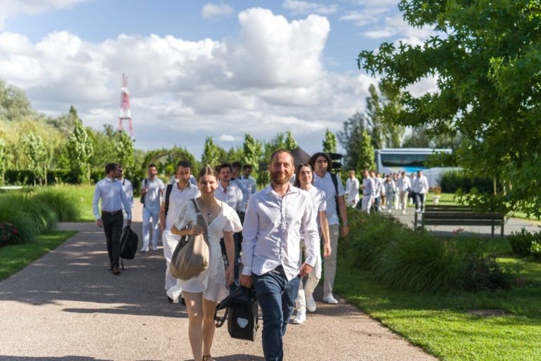 Un groupe de personnes vêtues de blanc et de couleurs claires marchent sur un chemin pavé à travers un parc verdoyant et luxuriant sous un ciel partiellement nuageux. Des arbres et de l'herbe entourent le chemin, avec une tour rayée rouge et blanche visible en arrière-plan. Un homme et une femme dirigent le groupe souriant, portant des sacs pour la Soirée annuelle du Groupe Betclic.