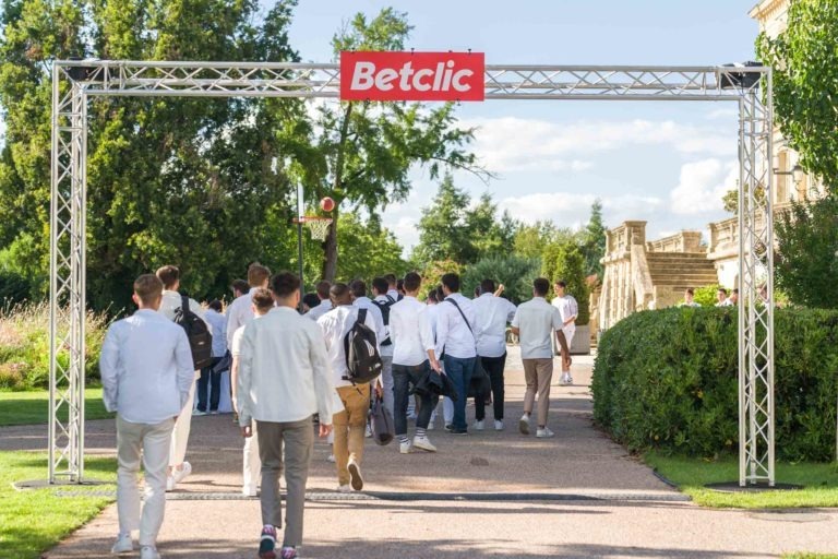 Un groupe de personnes, en majorité des hommes, s'éloigne de la caméra sous une arche métallique sur laquelle est inscrit « Betclic ». Ils se trouvent sur un chemin bordé de verdure luxuriante et de bâtiments historiques. La plupart d'entre eux portent des chemises blanches et des sacs à dos. La scène est lumineuse et ensoleillée pendant la Soirée annuelle du Groupe Betclic.