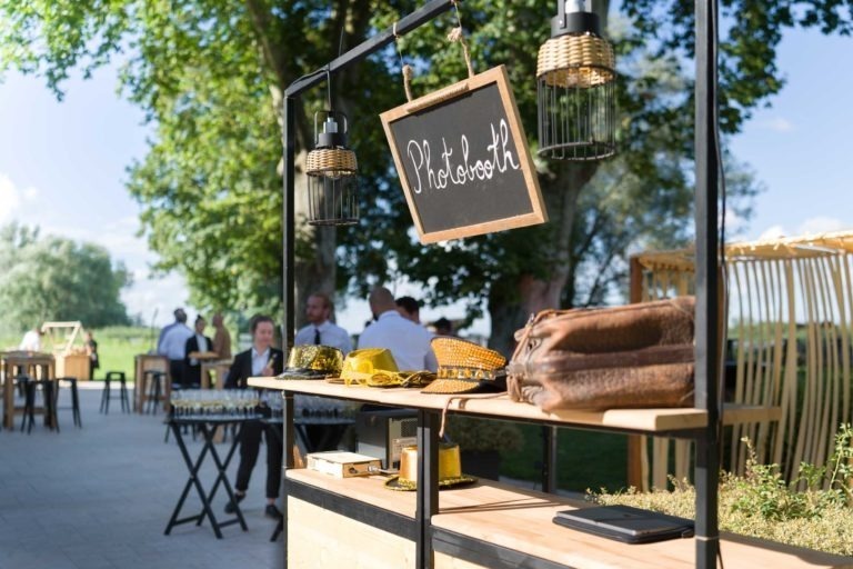 Un photomaton extérieur installé lors de la Soirée annuelle du Groupe Betclic comporte un panneau en ardoise sur lequel est écrit « Photobooth ». Des accessoires, notamment des chapeaux et des lunettes, sont disposés sur une table en bois. Des personnes en tenue de soirée sont à l'arrière-plan, et des arbres à la végétation luxuriante entourent la zone. La scène est ensoleillée et lumineuse.