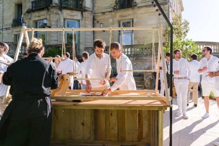 Un groupe de personnes vêtues de blanc se rassemble à l'extérieur, près d'un bâtiment historique aux balcons décorés. Lors de la soirée annuelle du groupe Betclic, deux hommes au premier plan préparent la nourriture derrière un comptoir en bois avec l'aide d'un individu vêtu de noir. D'autres discutent, la verdure et la lumière du soleil enrichissant la scène.