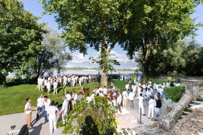 Un grand rassemblement extérieur pour la Soirée Annuelle du Groupe Betclic réunit de nombreuses personnes vêtues de blanc. Elles sont réparties sur une pelouse verte et une zone pavée entourée d'arbres abondants, avec un plan d'eau visible en arrière-plan. La scène évoque un événement social ensoleillé et détendu où les invités discutent.