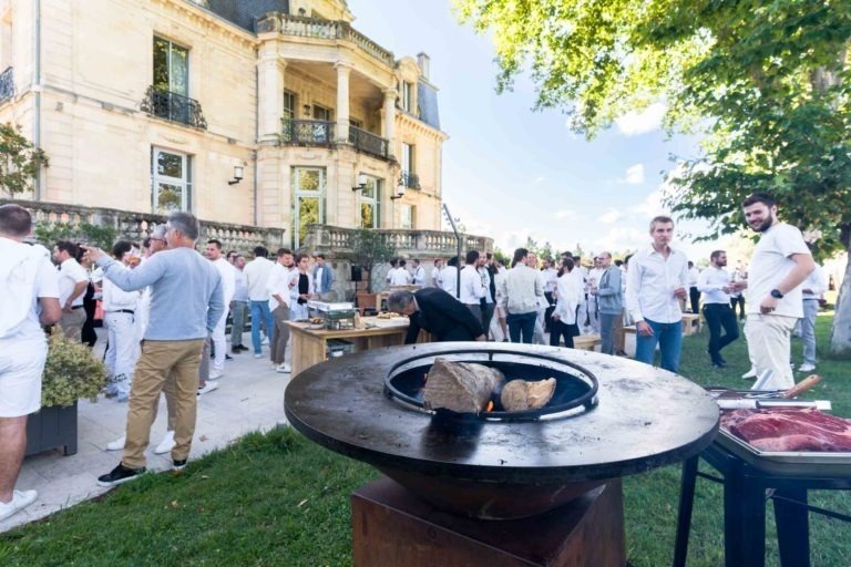 Un grand groupe de personnes vêtues de blanc se rassemblent devant un manoir historique pour la Soirée annuelle du Groupe Betclic. Au premier plan, un gril rond ouvert grésille et des aliments y cuisent. Certaines personnes discutent tandis que d'autres préparent à manger. La scène animée se déroule devant des arbres et un ciel bleu.