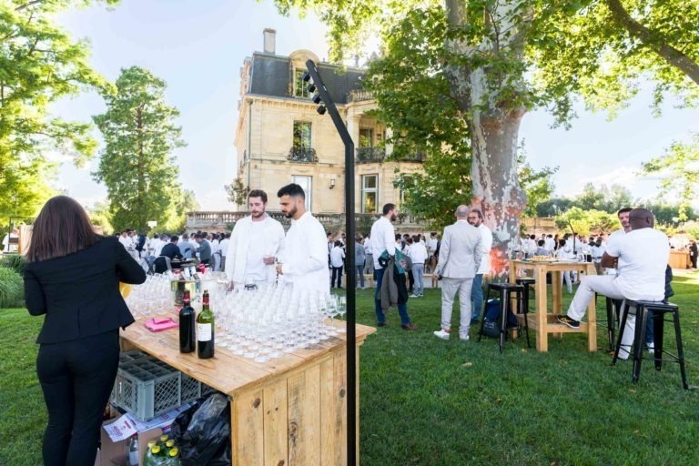 Des personnes vêtues de blanc sont rassemblées lors d'un événement en plein air, la Soirée annuelle du Groupe Betclic, devant un bâtiment historique. Il y a un bar en bois avec des bouteilles et des verres sur les deux tables et une femme prépare des boissons. Des tables de bar et des tabourets sont dispersés sur la pelouse verte, ombragée par de grands arbres.