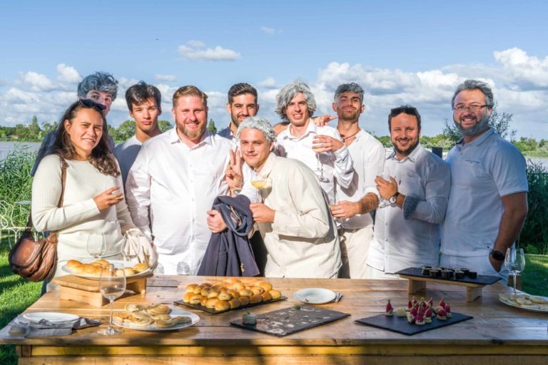 Un groupe de personnes, la plupart vêtues de blanc, posent à l'extérieur près d'un front de mer lors de la Soirée annuelle du Groupe Betclic. La table devant eux contient un assortiment de pâtisseries, de croissants et de verres de vin. Le ciel est bleu avec quelques nuages et de la verdure est visible en arrière-plan. Tous les individus sourient et semblent d'humeur festive.