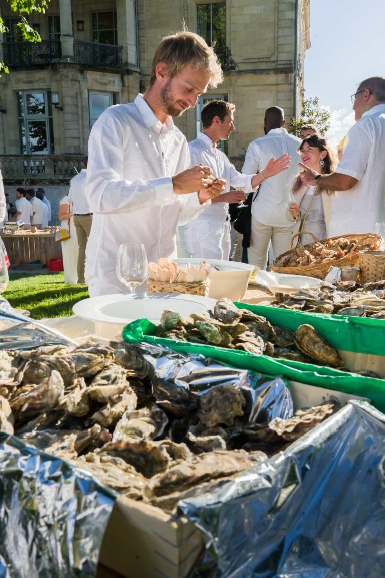 Un homme en chemise blanche ouvre des huîtres lors de la Soirée Annuelle du Groupe Betclic, un événement élégant en plein air. Il se tient près de tables remplies d'huîtres, tandis que d'autres participants en vêtements blancs discutent et profitent de l'occasion. L'événement se déroule dans un jardin luxuriant avec un grand bâtiment visible derrière les invités.