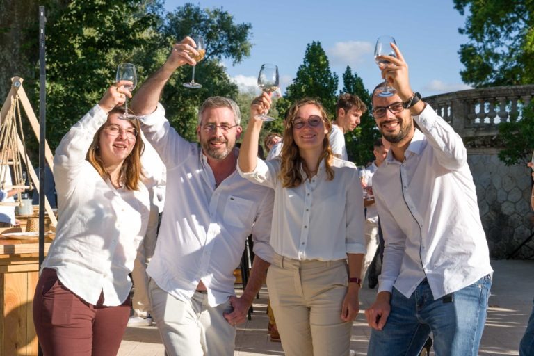 Quatre personnes se tiennent dehors dans un jardin, souriant et levant leurs verres de vin pour célébrer la Soirée annuelle du Groupe Betclic. Vêtus de chemises blanches, ils se tiennent près les uns des autres avec des arbres, un mur de pierre et un ciel bleu en arrière-plan. D'autres participants contribuent à l'atmosphère festive.