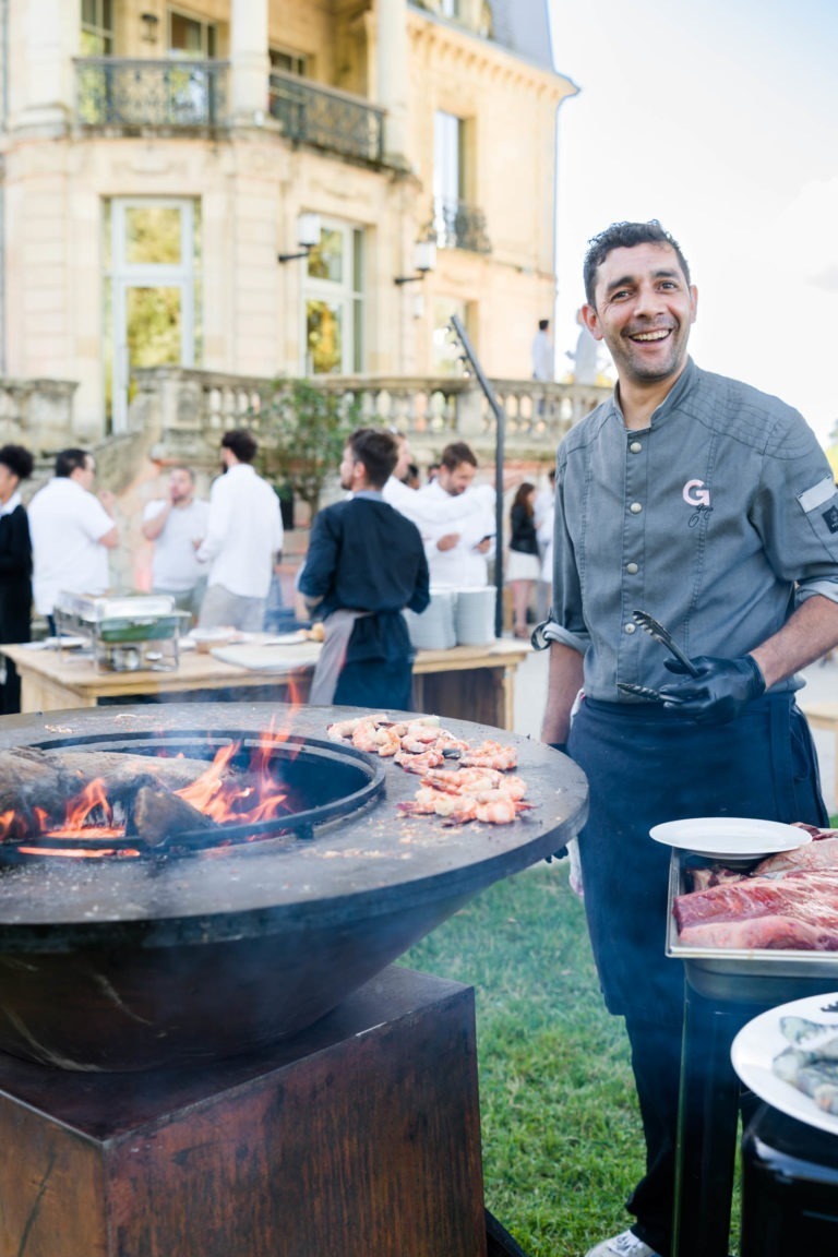 Un chef souriant se tient à côté d'un grand gril rond extérieur avec des flammes lors de la Soirée annuelle du Groupe Betclic. Des brochettes de nourriture cuisent sur le gril. Le chef porte un uniforme gris et des gants noirs. Derrière lui, un groupe de personnes est rassemblé près de tables avec de la nourriture, et un grand et élégant bâtiment est en arrière-plan.