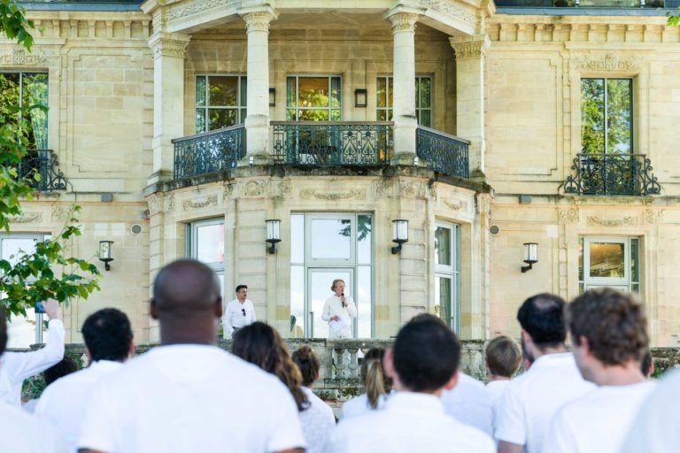 Un groupe de personnes vêtues de blanc fait face à un orateur debout sur les marches d'un grand bâtiment décoré avec de grandes fenêtres et un balcon. L'orateur est également vêtu de blanc et s'adresse à la foule à l'aide d'un microphone. Des arbres bordent le bâtiment et l'événement, qui fait partie de la Soirée annuelle du Groupe Betclic, se déroule en plein air à la lumière du jour.