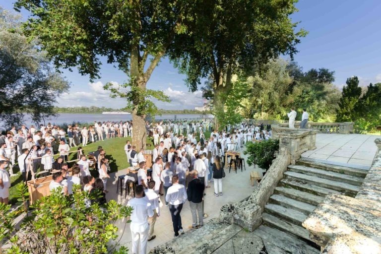 Un grand rassemblement en plein air, la Soirée annuelle du Groupe Betclic, réunit des gens vêtus de blanc au bord d'une rivière pittoresque. Les participants socialisent sous de grands arbres sur une herbe verte luxuriante, avec quelques individus près des marches de pierre et des balustrades au premier plan à droite. Le ciel bleu clair rehausse le cadre pittoresque.